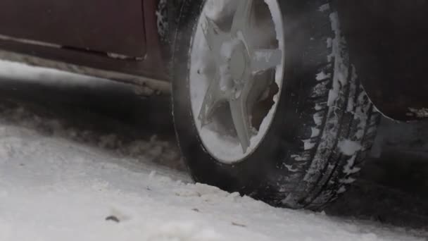 Araba kış şehrinde karla kaplı yolda duruyor. Egzoz borusundan duman gazları. Kar yağışı. Tekerleği yavaş çekimde kapat. Lastik. Trafik ve kar fırtınası için kötü hava koşulları. Yolculuk tehlikesi — Stok video