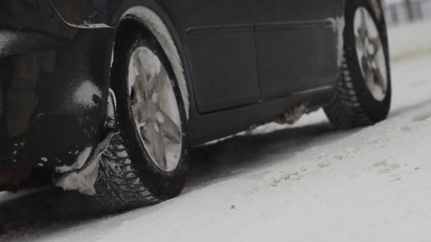 Samochód stoi na pokrytym śniegiem skrzyżowaniu w zimowym mieście. Gazy dymne z rury wydechowej. Śnieg. Zbliżenie koła w zwolnionym tempie. Opona. Złe warunki pogodowe dla ruchu, śnieżyca. Niebezpieczeństwo podróży — Wideo stockowe