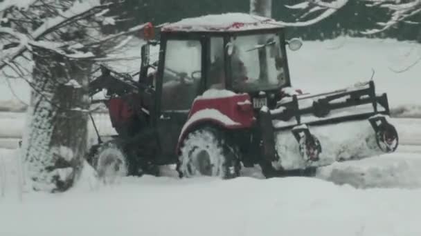 Een kleine trekker met een knipperend licht en een borstel rijdt op de weg om sneeuw te ruimen op de stoep na een sneeuwval — Stockvideo