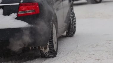 Araba kış şehrinde karla kaplı kavşakta duruyor ve dönüyor. Egzoz borusundan duman gazları. Kar yağışı. Tekerleği yavaş çekimde kapat. Lastik izleri. Kötü hava koşulları, kar fırtınası