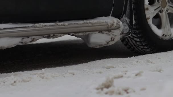 Autos stehen an Kreuzungen und setzen sich dann auf einer schneebedeckten Straße in winterlicher Stadt in Bewegung. Rauchgase aus dem Auspuff. Nahaufnahme eines Rades in Zeitlupe. Reifenprofil-Spuren. Schlechtes Wetter — Stockvideo