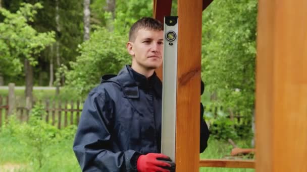 Un costruttore caucasico controlla la planarità della costruzione di un gazebo in legno fuori città in natura. Costruzione fai da te. Casa vacanza — Video Stock