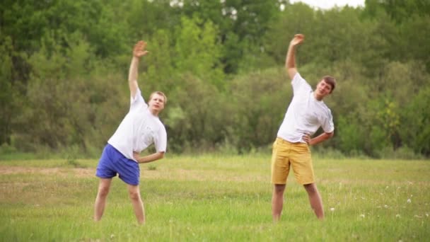 Dois jovens praticam esportes na natureza fazendo curvas de tronco. Atletismo na natureza, fundo — Vídeo de Stock