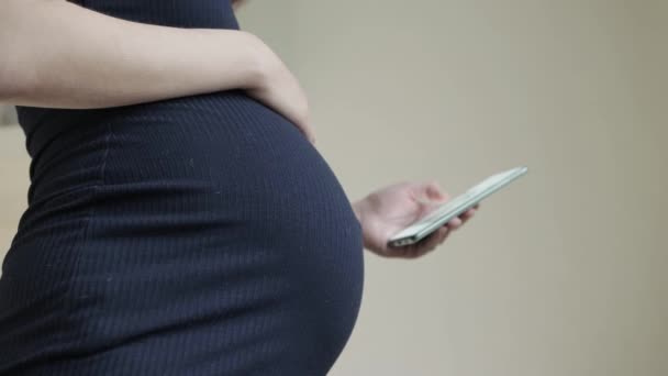Беременная девушка держит смартфон в руке на фоне живота. Концепция вреда гаджетов на развитие психики будущего здоровья ребенка, крупным планом — стоковое видео