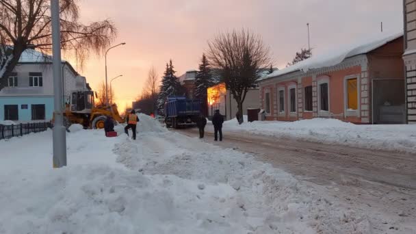 L'hiver. Tracteur balayeuse véhicule enlève la neige de la route à l'aide d'un seau. Coucher de soleil. La moisson des machines municipales nettoie la ville de la neige. Les travailleurs des services publics en gilet orange attendent le travail. — Video