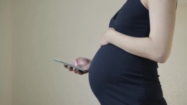 Hamile bir kız elinde bir akıllı telefon tutar karnının arka planına karşı. Çocukların gelecekteki sağlıklarının gelişimine yönelik cihazların zararları kavramı. Boşluğu kopyala