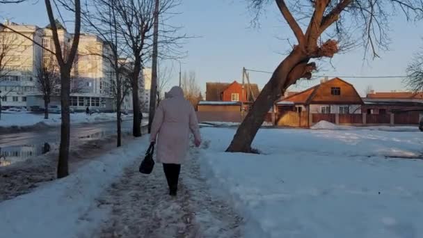 Mujer con chaqueta encapuchada camina a lo largo de un camino no despejado de nieve en una zona residencial de la ciudad en invierno. Primavera tras invierno. Temperatura por encima de cero. ¡Slush! Buenas tardes. Disparo de mano en movimiento — Vídeo de stock