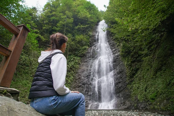Genç kadın bir şelale gözlemliyor, seyahat ederken doğanın tadını çıkarıyor.