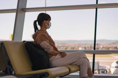 Yüz maskeli bir kadın havaalanında kalkış için telefonla konuşuyor. Dişi gezgin kalkış terminalinde bekliyor.