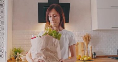 Elinde alışveriş poşeti olan kadın mutfakta durmuş kameraya bakıyor.