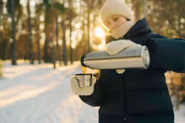Woman traveler holding thermos bottle and cup of tea, walking in snowy frozen winter forest. Adventure, travel, tourism and camping concept