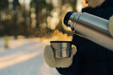 Kadın termostan bir fincana sıcak çay dökerek gün batımında karlı, donmuş kış ormanlarında yürür. Macera, turizm ve kamp konsepti