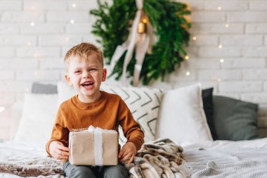 Noel sabahında hediyeleriyle gülen beyaz çocuk. Arka planda Noel ağacı çelengi var. Çocuk heyecanlı ve gülümsüyor. Mutlu aile tatilleri. Kutularda hediyeleri açmak. Boşluğu kopyala.