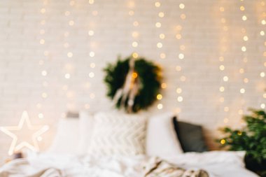 Kış tatili için muhteşem, davetkar, bulanık Noel yatak odası iç tasarımı. Yan tarafında Noel ağacı olan büyük, rahat bir yatak ve arka planında parlak, sıcak ışıklar olan büyük bir çelenk..