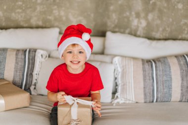 Kırmızı Noel Baba 'lı gülen beyaz çocuk Noel sabahı hediyelerini alıyor. Çocuk heyecanlı ve gülümsüyor. Hediyeleri açıyorum. Boşluğu kopyala.