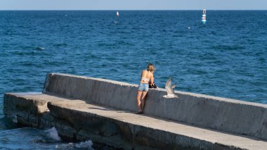 Bir kız mavi denizin arka planında beton bir iskelede duruyor, güneşli bir sonbaharın ilk günlerinde bir deniz kuşu yakınlarda uçuyor