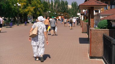 İnsanlar güneşli bir günde geniş bir set boyunca yürürler. Önde olgun bir kadın yürür.