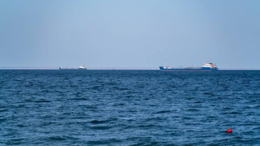 İki büyük nakliye gemisi, güneşli bir günde, deniz koyundaki yolda, gökyüzünün arka planına ve uzak kıyıların kenarına karşı dururlar.