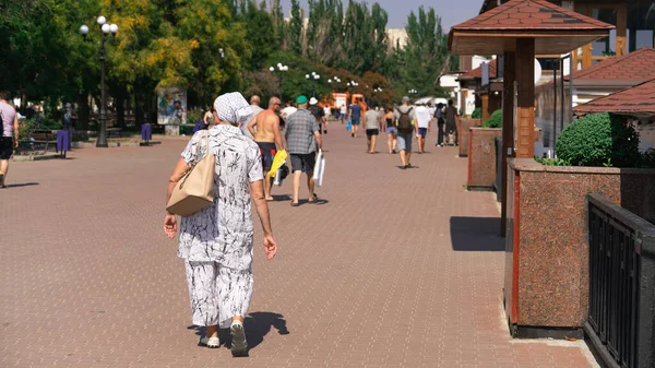 İnsanlar güneşli bir günde geniş bir set boyunca yürürler. Önde olgun bir kadın yürür.