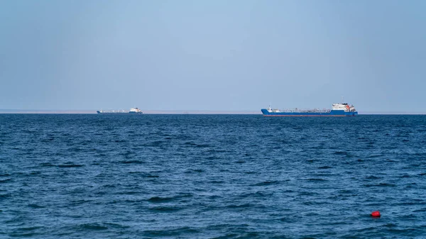 İki büyük nakliye gemisi, güneşli bir günde, deniz koyundaki yolda, gökyüzünün arka planına ve uzak kıyıların kenarına karşı dururlar.