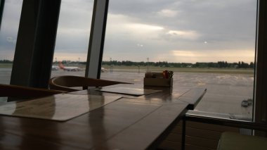Yaz sabahının erken saatlerinde havaalanı restoranında bir masa. Arka planda, uçakların siluetleri geniş bir pencereden görülebiliyor.