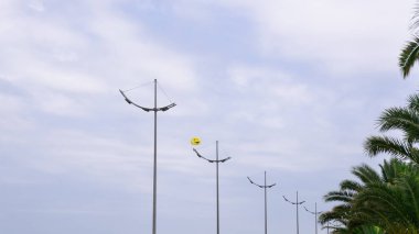 Neşeli bir gülümseme şeklinde sarı bir paraşüt, mavi bulutlu gökyüzüne karşı tekneler şeklindeki lambalarla uzaktaki elektrik direklerinin üzerinde asılı duruyor.