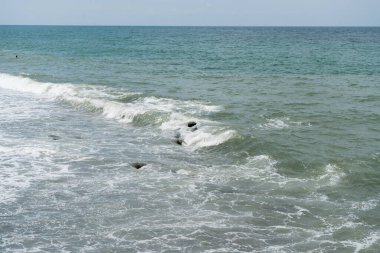 Deniz dalgaları kıyıların kıyısında güneş ışınlarıyla köpürüyor, kıyı taşlarını açığa çıkarıyor, uzaktan banyo yapan bir adamın başını görebilirsiniz.