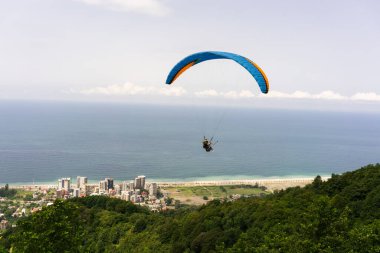 İki paraşütçü dağın üzerinden uçuyor ve mavi denize doğru evler ve güneşli bir yaz gününde ufukta beyaz bulutların çizgisi.