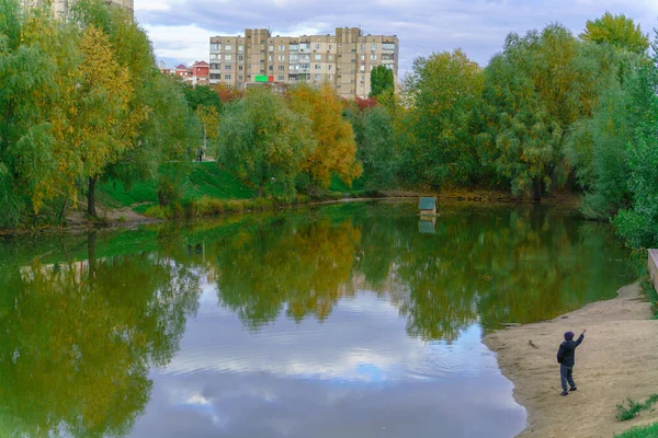 A boy in an autumn evening throws stones into a lake, on the surface of which autumn trees and the sky are reflected in a city park