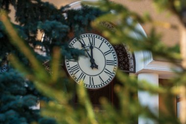 Siyah saat büyük beyaz bir kadranda 5-12 arası caddedeki yeşil çam dallarının arasından geçen bir evin duvarında.