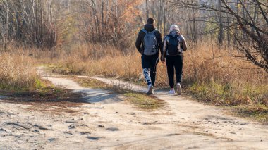 Arkalarında sırt çantası olan genç bir adam ve bir kız güneşli bir günde yapraksız ağaçlara giden toprak yolda el ele yürürler.