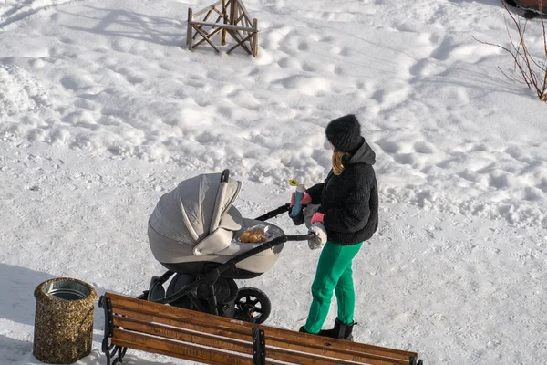 Genç bir anne termostan sıcak bir içecek içer ve yabancılardan uzakta, güneşli bir kış gününde bebek arabasının yanında kurabiye yer.