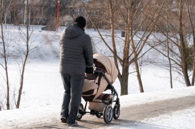 Genç bir baba, güneşli bir kış gününde bir bebek arabasıyla şehir parkında yürüyor.