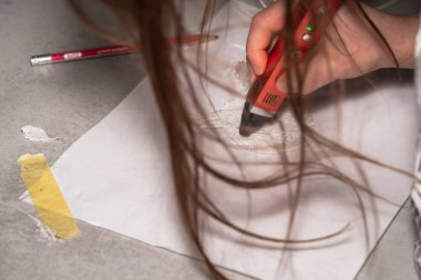 Gevşek saçlarının arkasında, küçük bir kızın çocuksu eli kırmızı bir 3D yazıcı kalemi tutuyor ve beyaz bir kağıt üzerinde beyaz iplikten paskalya tavşanı yapıyor. Üst taraf görünümü