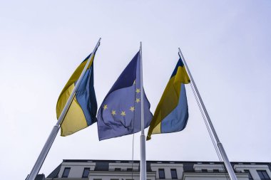 Avrupa Birliği bayrağı ve sağda rüzgarda dalgalanan iki Ukrayna bayrağı binanın ve mavi gökyüzünün arka planına karşı. Alt kenar görünümü