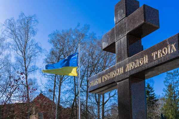 "God save your people" - written on a granite cross next to the flag of Ukraine, fluttering on a metal flagpole in the wind against the backdrop of trees and a blue sky