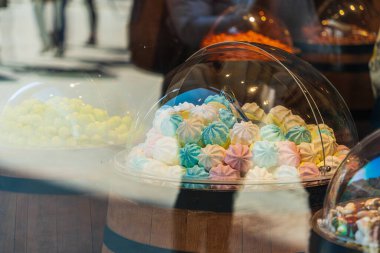 Güneşli bir günde yoldan geçenlerin camlarında yansımaları olan bir pastanenin vitrininin arkasında çok renkli tatlı kekler ve marşmelovlar.