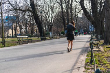 Kısa deri ceketli ve etekli yalnız bir kız, omzunda bir çantayla şehrin parkında asfalt bir yol boyunca yürür ilkbaharın başlarında güneş ışığına doğru. Arka plan