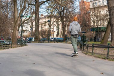 Genç bir adam, ilkbaharın başlarında şehir meydanında asfalt bir yolda snowboard sürüyor. Arkadan bak.