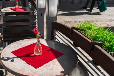Krakow 'da bir sokak kafesinin gri yuvarlak masasında kırmızı bir peçetenin üzerindeki cam bir vazoda beş kırmızı karanfil.