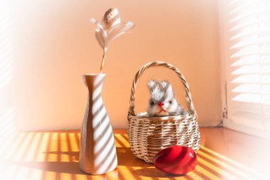 Turuncu bir yüzey üzerindeki mutlu bir Paskalya bayramının özellikleri. Panjurlardan süzülen ışık çizgileri. Turuncu bir yumurta, sepette bir tavşan, beyaz vazoda bir yumurta.