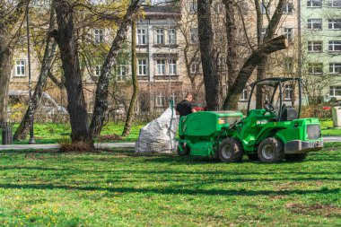Yeşil çimenleri temizlemek için bir belediye aracının sürücüsü şehir parkındaki yeşil bir çimenlikteki hasat makinesi ve çöp toplama çantasının yanında duruyor. Güneşli gün erken bahar