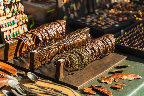 Takma metal bileklikler, broşlar, el yapımı demirci kaşıkları ve çatalları bir tezgahın üzerinde güneş ışığında ahşap bir standın üzerinde parlıyor..