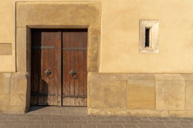 Ortaçağ ahşap kapısı, yuvarlak demir kulpları ve klasik duvardaki dar penceresi akşam güneşinde taş kaldırım. Doğal tarihi geçmiş