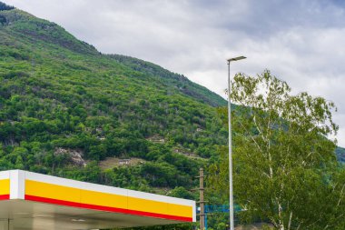 Street lighting pole, bright roof of a gas station, boom of a construction crane near a mountainside with a green forest in the Swiss Alps, civilization and nature