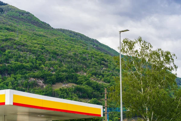 Sokak aydınlatma direği, bir benzin istasyonunun parlak çatısı, İsviçre Alpleri, medeniyeti ve doğasında yeşil bir ormanı olan bir dağ yamacının yakınındaki bir vinç patlaması.