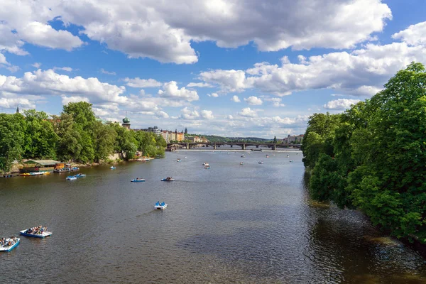 Güneşli bir günde Prag, Çek Cumhuriyeti 'ndeki Vltava nehrinde, beyaz bulutlar ve kıyı şeridinde uzun yeşil ağaçlar bulunan mavi bir gökyüzünün altında, tatilcilerle birlikte sayısız zevk teknesi, Avrupa turizmi