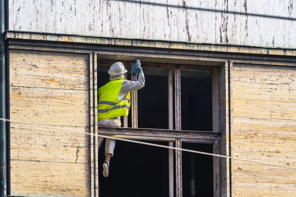A woman in the form of a builder on a sunny day at a height with a special tool grinds the wooden frame of the window of an old house during its repair, reconstruction of old houses in Prague