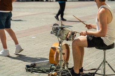 Bir sokak müzisyeni, Avrupa 'da güneşli bir günde gri bir meydanda beyaz tişört ve siyah şortla perküsyon enstrümanları çalıyor. Yan görünümü kapat