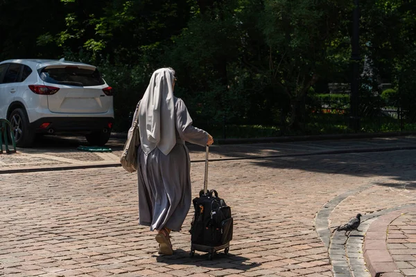 Yaşlı Polonyalı bir rahibe, tekerlekli bir bavul ve omuzunda bir çanta ile, asfalt bir yolda yürür parlak gün ışığında karanlığa, önünde bir güvercin ona yol açar.